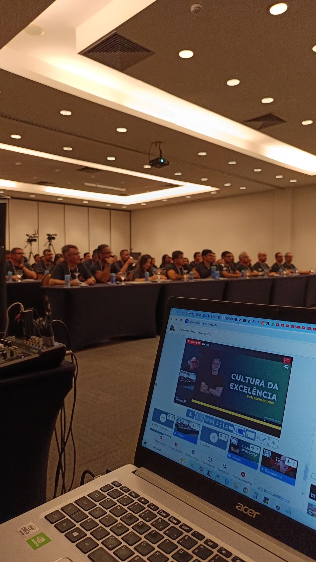 Público de evento presencial e operação de transmissão ao vivo em notebook